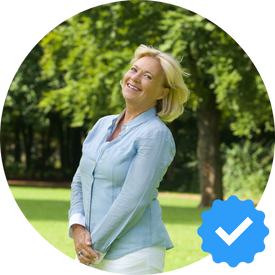 A happy woman with blonde hair, standing outdoors in a park, smiling, and wearing a light blue blouse. A verification badge icon is visible in the lower right corner.