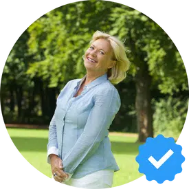 A happy woman with blonde hair, standing outdoors in a park, smiling, and wearing a light blue blouse. A verification badge icon is visible in the lower right corner.