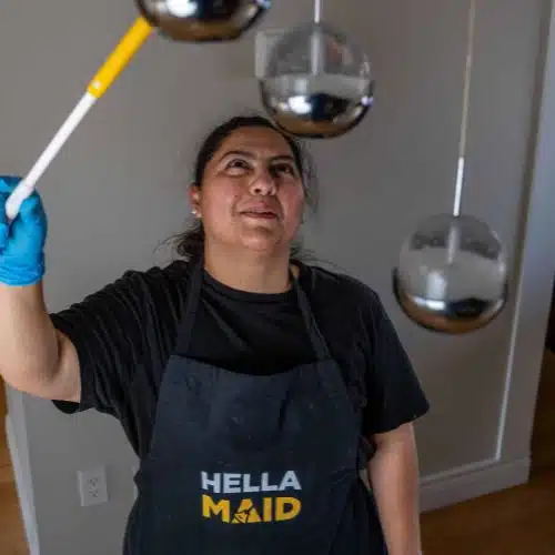 A professional cleaner wearing a 'Hellamaid' apron and gloves smiles while dusting modern hanging light fixtures with an extendable duster in a well-lit home