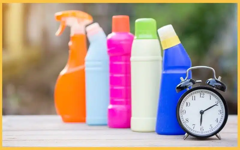 Five colorful plastic cleaning bottles lined up on a table beside an analog clock, emphasizing why Canadians book professional cleaning to save time.