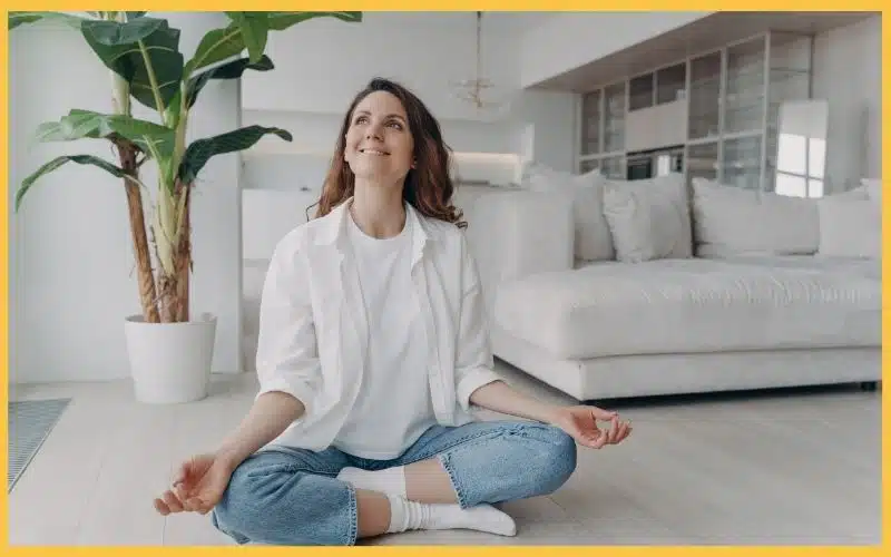 A woman sits cross-legged on floor in a clean modern living room, meditating peacefully.