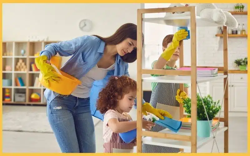 An adult and children clean shelves using various cleaning supplies while wearing gloves, like when booking professional cleaning services do.