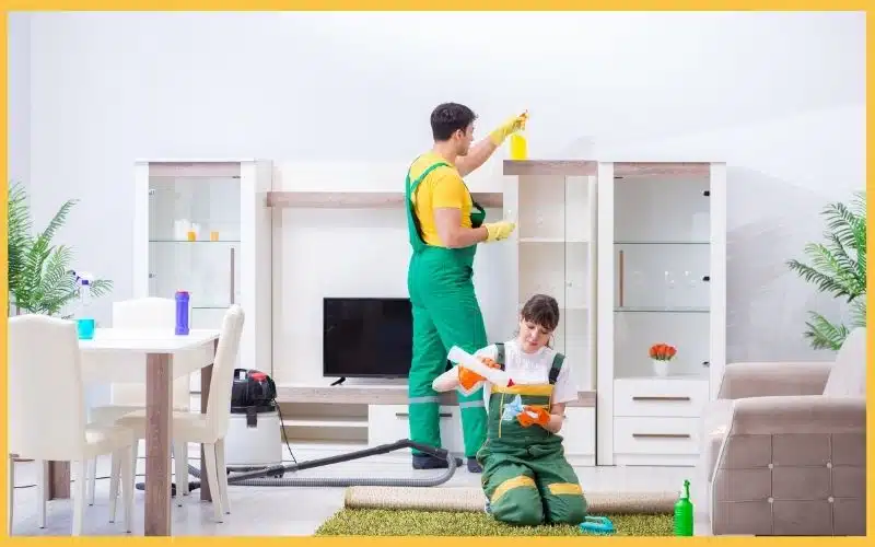 Two professional cleaners clean a living room, one applying cleaner to a shelf and the other scrubbing the floor.