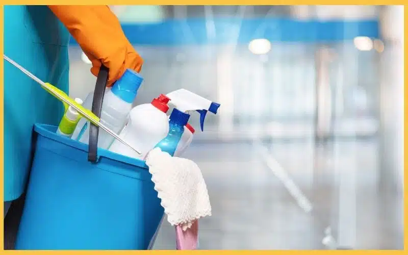 A person in orange gloves holds a blue bucket filled with cleaning supplies, including spray bottles and a microfiber cloth, symbolizing hiring a cleaning company for your needs.