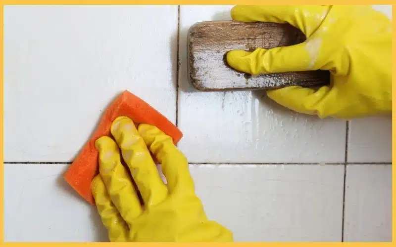 Close-up of hands in yellow gloves cleaning tiled surfaces with an orange sponge and a wooden scrubbing tool to symbolizing the cleaning companies near me question.