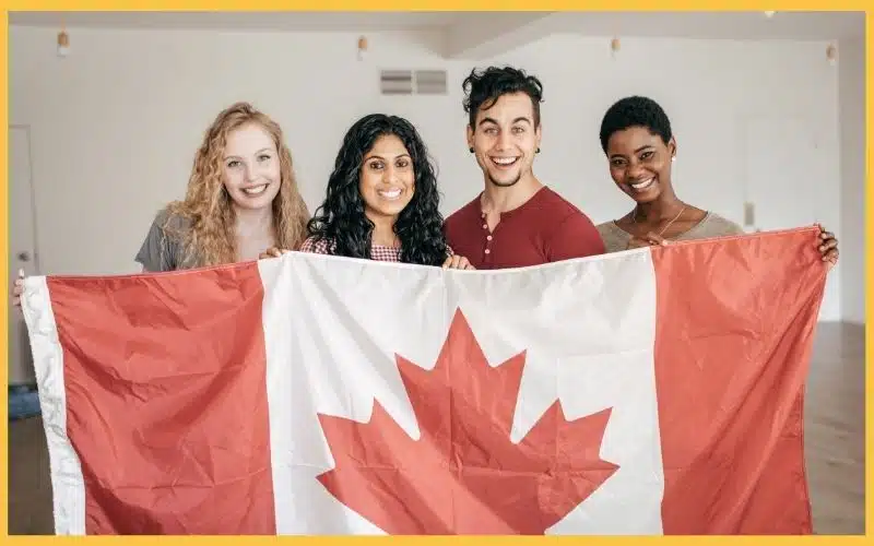 A diverse group of four people proudly holding a large Canadian flag, posing in a clean and spacious room.