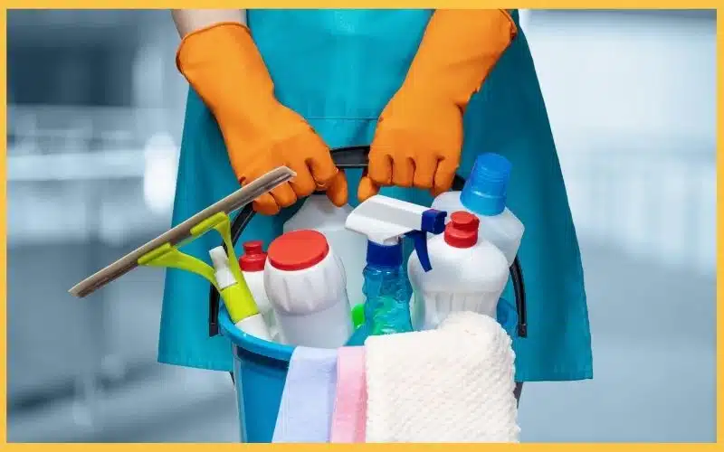 A person in orange gloves holds a cleaning bucket filled with various bottles, sprays, and a towel, ready for cleaning tasks.