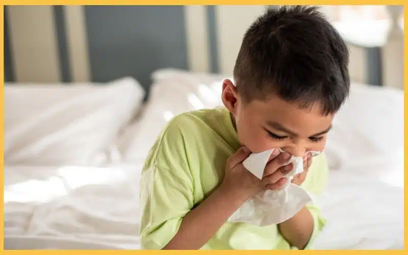A child wearing a bright green shirt is sitting on a bed, holding a tissue close to their face, in a softly lit room.