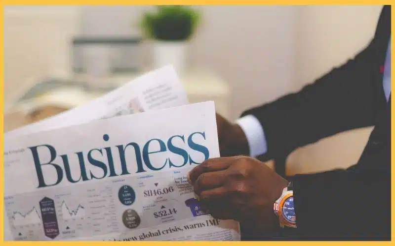 A man in a suit holds a business newspaper featuring market data and graphs, seated in a bright office setting.