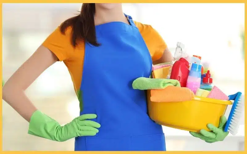 A woman in a blue apron and green gloves holds a yellow bucket filled with cleaning supplies and colorful towels, ready to clean.