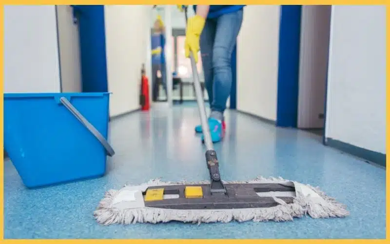 A person with yellow gloves mops a blue-tiled hallway, with a bucket nearby, indicating a cleaning task in progress.