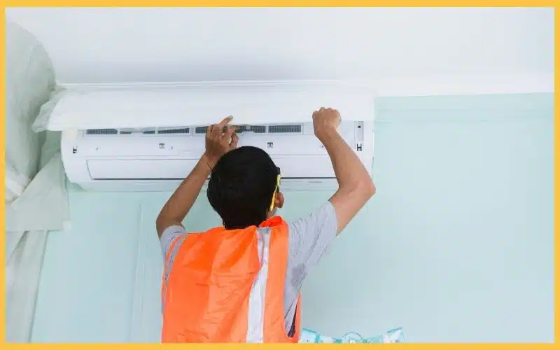 A technician in an orange safety vest installs an air conditioner on a light blue wall, focused on the unit above.
