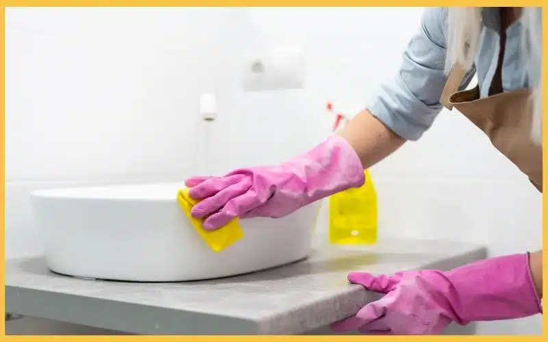 A person wearing pink gloves cleans a white sink with a yellow sponge on a gray countertop, with cleaning supplies nearby.