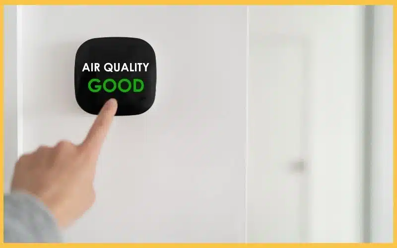 A hand points to a wall-mounted air quality monitor displaying "AIR QUALITY GOOD" in bold green letters.