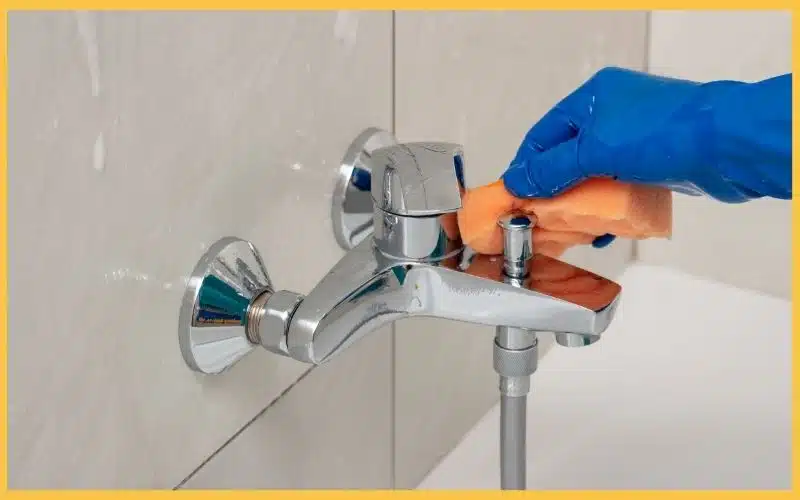 A gloved hand cleans a chrome faucet with a sponge, droplets visible on the tiled wall behind.