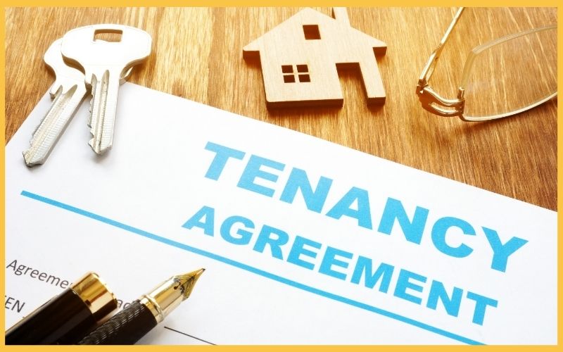 A tenancy agreement document with blue text, keys, a wooden house model, and reading glasses on a wooden surface.