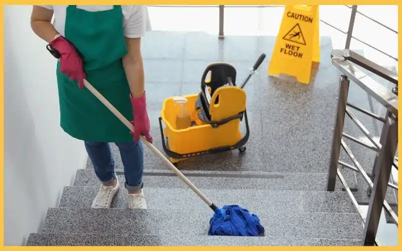 A worker in pink gloves mops a stairway, with a yellow bucket and a caution sign for a wet floor in the background.
