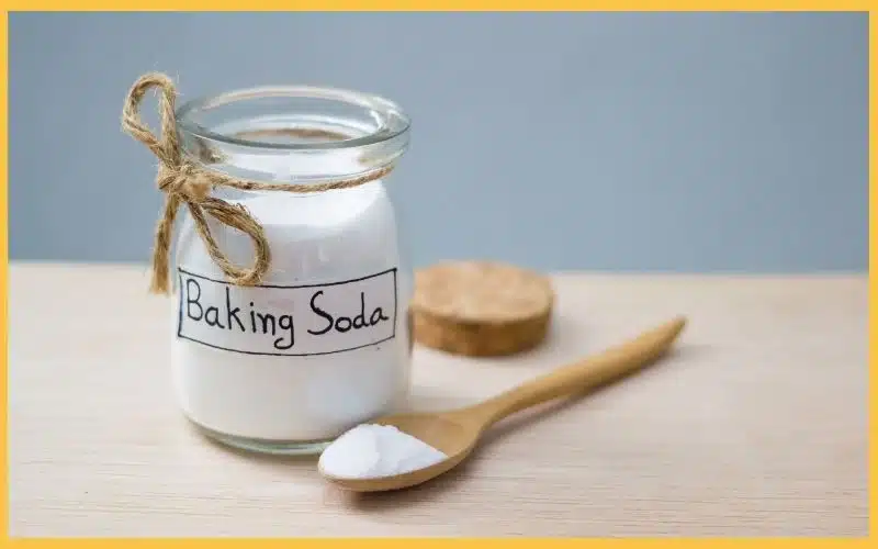 Glass jar labeled "Baking Soda" with twine, next to a wooden spoon with a small heap of baking soda on a light wooden surface.