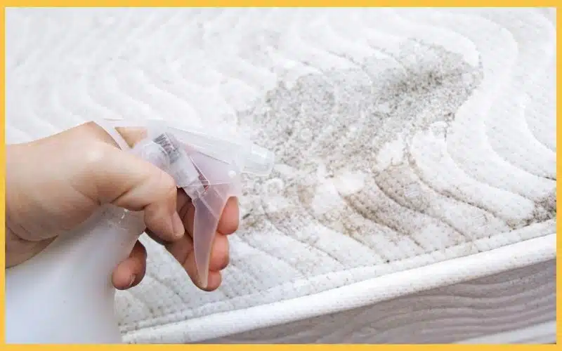 A hand holding a spray bottle, cleaning a stained mattress surface with visible dirt and discoloration.