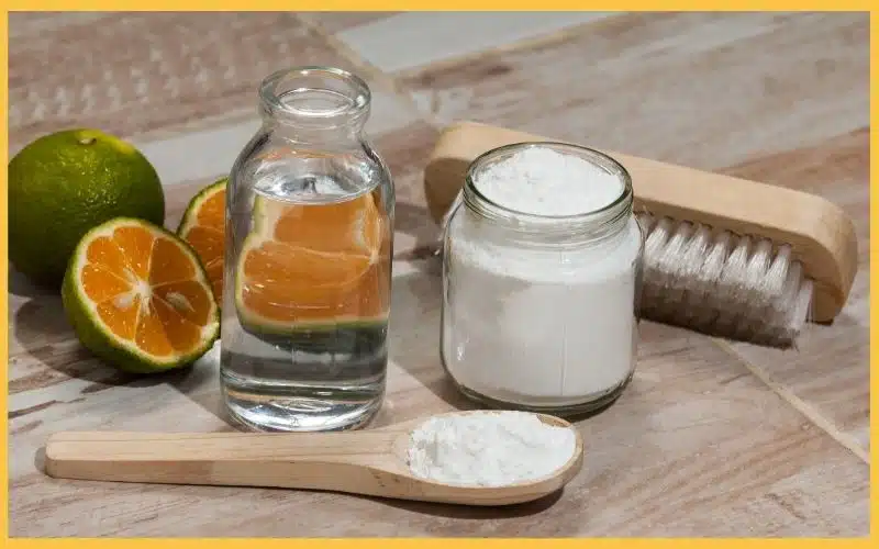 A bottle of water, a jar of white powder, and a wooden spoon next to halved green limes and a scrub brush on a tiled surface.