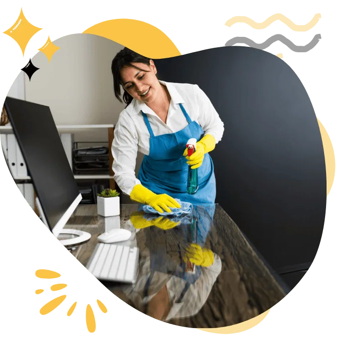 Professional office cleaner in an apron and yellow gloves cleaning a desk with spray and cloth, showcasing Hellamaid's commercial cleaning services for a spotless workplace.