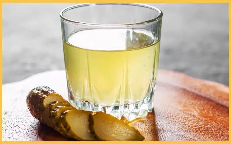 A glass of light yellow pickle juice next to sliced pickles on a wooden surface.