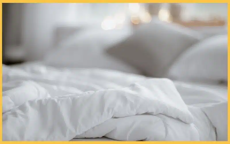 Close-up of a neatly arranged white comforter on a bed, with a soft gray pillow in the background and warm lights softly glowing.