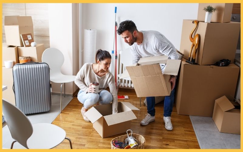 A couple is unpacking boxes in a cozy, cluttered room filled with moving supplies and a suitcase, preparing to settle in.