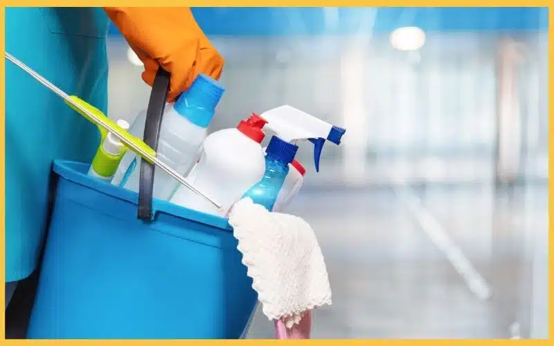 A gloved hand of a cleaning professional holding a bucket filled with cleaning supplies.