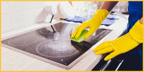 Gloved hands using a sponge to clean a kitchen's stove surface.