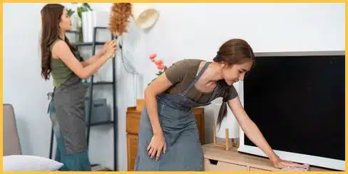 Two people cleaning the living area; one is dusting, and the other is wiping the surface.